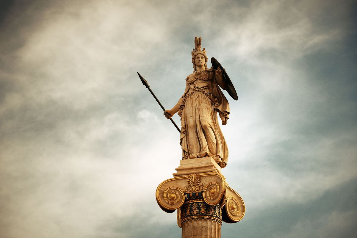 The image shows a statue of the goddess Athena standing atop a tall, ornate column. Athena is depicted in full armor, holding a spear and shield, set against a cloudy sky.